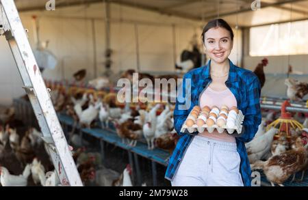 Farmerin in kariertem Hemd, hält ein spezielles Tablett mit aus biologischem Anbau geernteten Hühnereiern und lächelt vor der Kamera im Stall Stockfoto