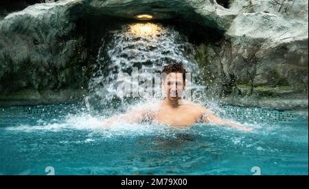 Der junge Mann genießt das planschende, fallende Wasser unter dem Wasserfall im Wellness-Pool des Holiday Spa, Kopierbereich Stockfoto