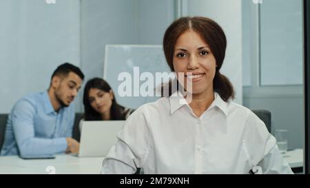 Porträt lächelnde, attraktive Geschäftsfrau, die in einem modernen Geschäftsbüro mit einem Team aus multikulturellen Profis sitzt und in die Kamera schaut. Freundliche Std Stockfoto
