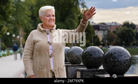 Porträt einer lächelnden, gealterten schönen Frau, die wegschaut, winkt hallo oder Auf Wiedersehen, die sich bei kühlem Wetter im Freien entspannt, eine glückliche ältere, grauhaarige Frau Stockfoto