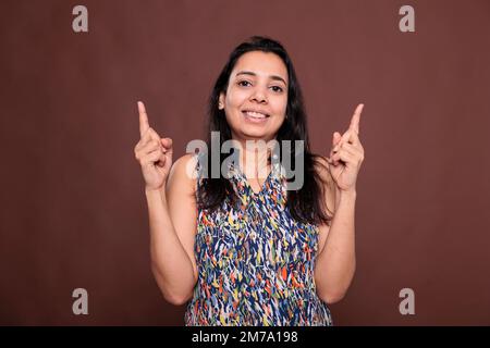 Lächelnde indianerin, die nach oben zeigt, mit Zeigefinger, Porträt, Werbeartikel. Fröhliche Dame steht, posiert, schaut in die Kamera, zeigt Beförderung, Studio Medium Shot Stockfoto