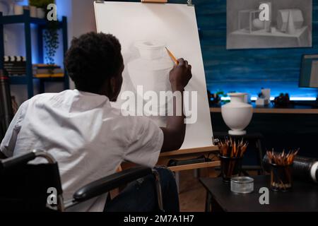 Männlicher Zeichner, der mit Gehbehinderung lebt und Bleistiftkunst von Topf kreiert. Ein afroamerikanischer Mann mit künstlerischen Fähigkeiten, der im Rollstuhl unterwegs ist, um Bilder auf Leinwand zu malen. Stockfoto