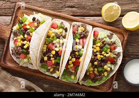 Mehltortillas gefüllt mit Hackfleisch und Gemüse, Salat, Tomaten, Mais, grüner Paprika und Bohnen, serviert auf Holzbrett Stockfoto