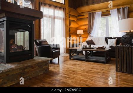 Propan-Gas-Glas-Kamin auf erhöhtem Flagstone-Sockel, brauner Ledersessel, hölzerner Couchtisch und Sofa im Wohnzimmer im luxuriösen Blockhaus. Stockfoto