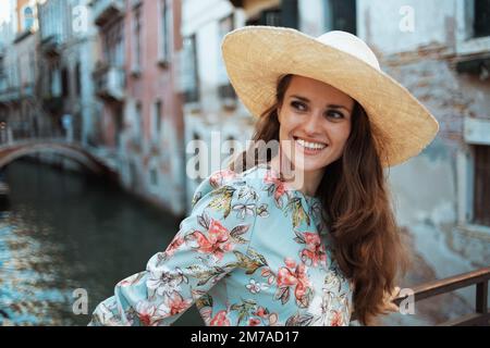 Eine glückliche, moderne Alleinreisende in Blumenkleid mit Hut genießt die Promenade in Venedig, Italien. Stockfoto