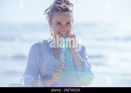 Porträt einer lächelnden stilvollen Frau mit einem Drink am Strand. Stockfoto