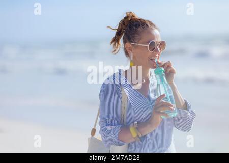 Lächelnde elegante 40-jährige Frau mit weißem Strohbeutel und Getränk am Strand. Stockfoto