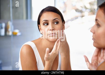 Ist das ein Fleck? Eine schöne junge Frau, die ihr Gesicht im Spiegel sieht. Stockfoto