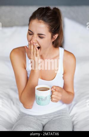 Immer noch so schläfrig... Eine junge Frau gähnt, während sie mit einer Tasse Kaffee im Bett sitzt. Stockfoto