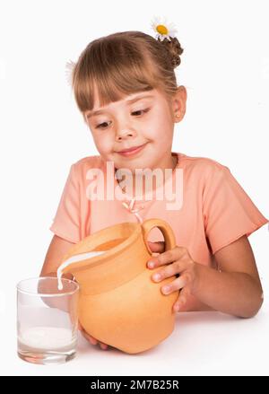 Gesundes Essen. Ein kleines Mädchen in einem orangefarbenen T-Shirt und Blumen im Haar gießt frische Milch aus einer Kanne auf weißem Hintergrund ein Stockfoto