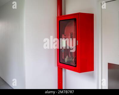 An der Wand in der Ecke des weißen Hotelgebäudes montierter Feuerlöscherschrank. Verschlossener roter Notfallkasten mit Feuerlöscher und Feuerlöschschlauch Stockfoto