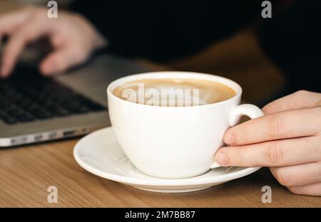 Nahaufnahme, eine Tasse Kaffee und auf einem unscharfen Hintergrund mit einem Notebook. Stockfoto