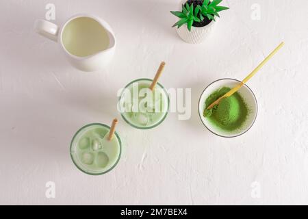 Zwei Gläser mit grünem Eistee Matcha Latte, eine Schüssel Matcha Puder, ein Milchkännchen mit Milch. Draufsicht. Weißer Hintergrund wird gekocht. Wellness-Drink Stockfoto