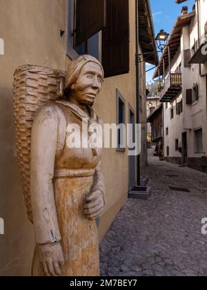 Sutrio, Italien - 29. Dezember 2022: Lebensgroße Holzskulptur einer Frau mit einem Korb aus Rucksack-Korb auf der Straße eines Dorfes Sutrio Stockfoto