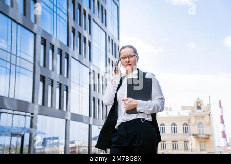 Erfolgreiche Geschäftsfrau oder Managerin, Frau, die mit dem Mobiltelefon unterwegs ist und ihr Notebook in der Hand hält. Die Geschäftsfrau der Stadt arbeitet in Anzug und Brille. Stockfoto