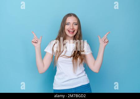 Hey, sieh mal. Lächelndes, wunderschönes weibliches Model, das mit dem Finger auf einen leeren Raum zeigt, und es dazu einlädt, sich eine Anzeige auf blauem Hintergrund anzuschauen. Tragen weißes T-Shirt und Mütze als Mockup Stockfoto