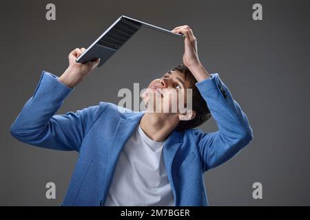 Ein glücklicher, weißer junger Kerl in blauer Jacke, der einen Laptop über den Kopf hält, isoliert auf grauem Hintergrund. Stockfoto