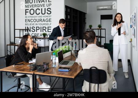 Frau, die in einem stilvollen Büro vor verschiedenen nationalen Mitarbeitern einen Bericht über Zeit-Brainstorming abgibt Stockfoto