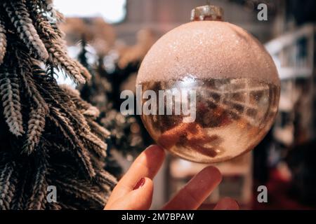 Große goldpinke Glaskugel in Frauenhand auf verschwommenem Hintergrund. Hintergrund für Weihnachtsbaumspielzeug. Inneneinrichtung für Silvester 2023. Ich habe mich entschieden Stockfoto