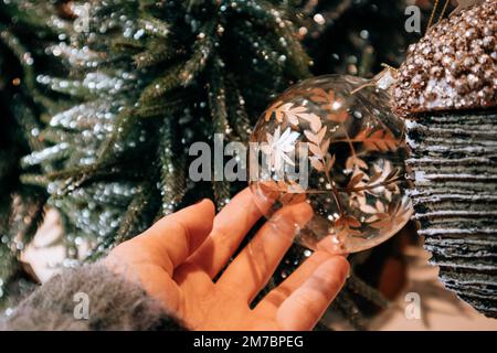 Weihnachtsspielzeug. Große goldene Glaskugel in der Hand einer Frau auf Baumhintergrund im neuen Jahr. Inneneinrichtung für Silvester 2023. Spielzeuge für jeden Geschmack auswählen Stockfoto