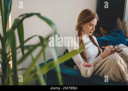 Junge rothaarige Frau schreibt Tagebuch in einem gemütlichen Haus auf dem Sofa im Wohnzimmer. Entspannungs-Lifestyle-Konzept. Stockfoto