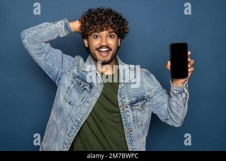 Modell, Kopierbereich. Ein fröhlicher, aufgeregter indianer oder araber hält in legerer Kleidung ein Smartphone mit leerem weißen Bildschirm zur Präsentation und zeigt mit dem Finger Stockfoto