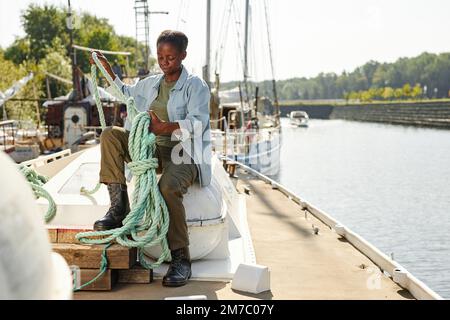 Ein langes Porträt einer jungen schwarzen Frau, die ein Seil an Bord in Docks bindet, beleuchtet von Sonnenlicht, Kopierraum Stockfoto