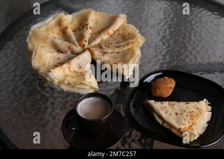 Foto von leckeren gebratenen Pfannkuchen und abgekochter Kondensmilch auf einem Teller und einer Tasse Kaffee auf einem Glastisch Stockfoto