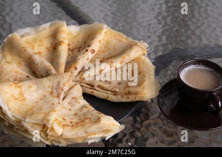 Foto einer Gruppe dreieckiger Pfannkuchen, die in einem Kreis auf einem Teller gestapelt sind, und einer Tasse Kaffee auf einem Glastisch Stockfoto