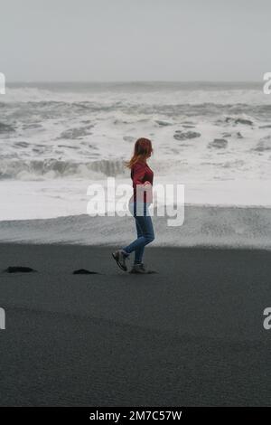 Frau, die am schwarzen isländischen Strand entlang spaziert und malerische Fotos macht Stockfoto