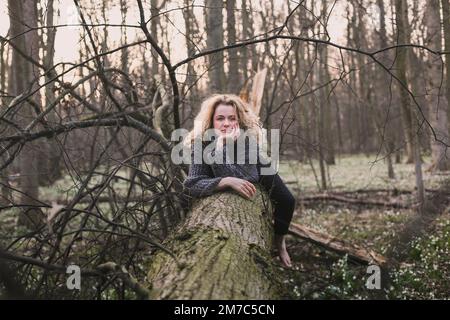 Blonde Frau, die auf einem großen gefallenen Baum sitzt, malerische Fotografie Stockfoto