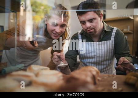 Ein Mann mit Down syndrom, der seinem Kollegen in einem Café hilft. Konzept der gesellschaftlichen Integration von Menschen mit Behinderungen. Stockfoto