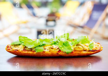 Baguette mit Tomatensauce, Fleisch, Jalapeno und Rucola, gebacken mit Käse auf einem Tisch im Freien. Beliebtes Street Fast Food Stockfoto