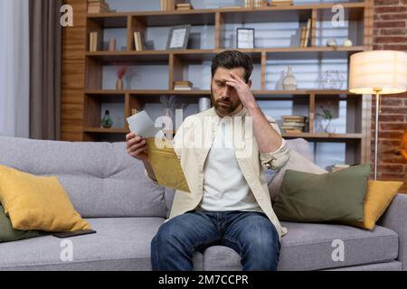 Verärgerter Mann zu Hause, der auf dem Sofa sitzt und einen Brief liest, mit schlechter Nachricht in Umschlagöffnung, unglücklich im Wohnzimmer und traurig. Stockfoto