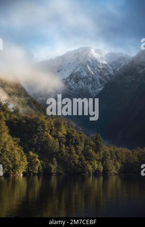 Ein magischer Morgen im Doubtful Sound, wenn frischer Schnee die Gipfel aus dem Wasser streicht, umgeben von niedrigen Wolken. Stockfoto