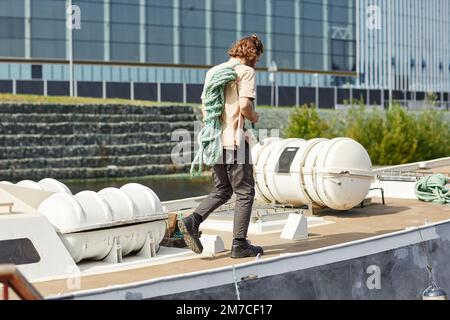 Rückansicht eines langhaarigen jungen Mannes, der auf einem Boot läuft und ein Seil im Sonnenlicht hält, Kopierraum Stockfoto