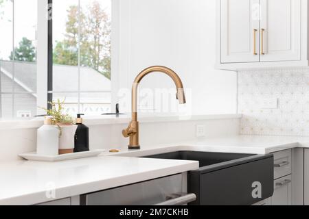 Ein wunderschönes Waschbecken in einer renovierten modernen Bauernhausküche mit goldenem Wasserhahn, schwarzem Bauernhaus-Waschbecken, weißem Granit und gefliestem Backplash. Keine Etiketten. Stockfoto