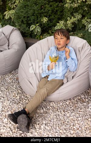Porträt eines kleinen Jungen, der Saft in einem Glas trinkt, dekoriert mit Obst, mit Stroh im Außenpark. Kind im Sommer Stockfoto