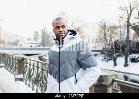 Junger afrikanischer Mann in legerer weißer und blauer Kleidung, spaziert am Fluss entlang. Straßenporträt mit Schnee in der Wintersaison. Lifestyle, Stadtleben Konzept Stockfoto