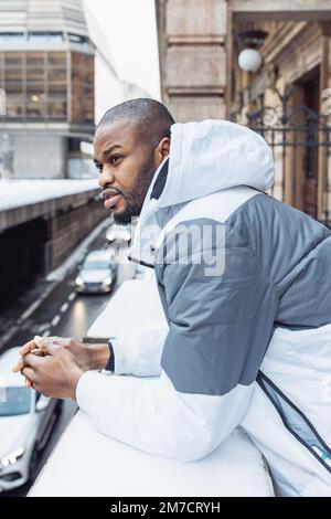 Selbstbewusster junger afrikanischer Mann in legerer weißer und blauer Kleidung, gestreifter Pullover. Straßenporträt mit Schnee in der Wintersaison. Lifestyle, Stadtleben Konzept. Seitenansicht Stockfoto