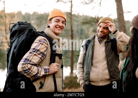 Seitliches Porträt eines lächelnden jungen Mannes mit Rucksack, der bei Freunden steht Stockfoto