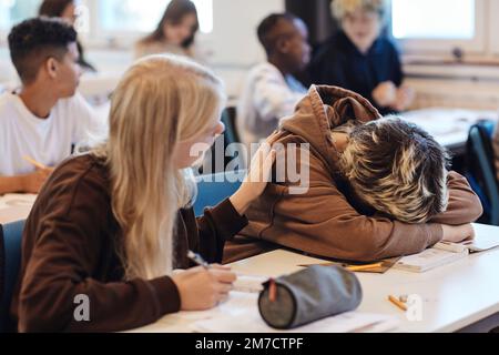 Blondes Teenager Mädchen tröstet eine deprimierte Freundin, die ihren Kopf im Klassenzimmer auf dem Schreibtisch ruht Stockfoto