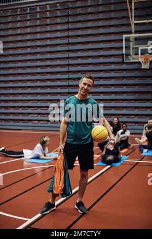 Das ganze Porträt eines lächelnden männlichen Trainers, der mit Basketball gegen Schüler auf dem Sportplatz steht Stockfoto