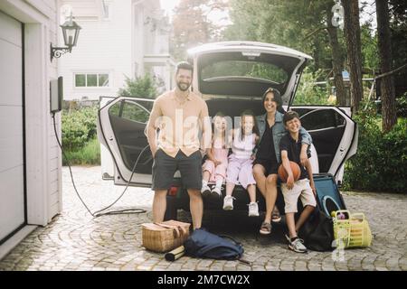 Porträt einer glücklichen Familie, die vor dem Haus im Elektroauto-Kofferraum sitzt Stockfoto