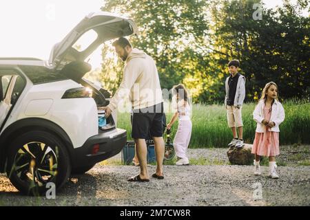 Vater entlädt Sachen aus dem Kofferraum eines Elektroautos auf einer unbefestigten Straße Stockfoto