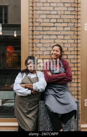 Porträt von lächelnden weiblichen Cafetbesitzern, die Schürzen tragen und die Arme vor dem Café kreuzen Stockfoto