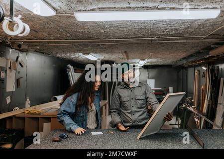 Ein erfahrener Handwerker zeigt der Kundin in der Werkstatt einen Bilderrahmen Stockfoto