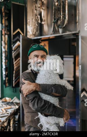 Porträt eines lächelnden Handwerkers, der seinen Hund in einem Rahmenladen umarmt Stockfoto