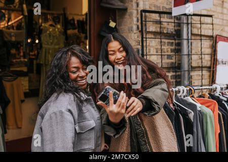 Fröhliche Freundinnen machen Selfie über Smartphone, während sie sich im Kleiderladen draußen Vergnügen Stockfoto