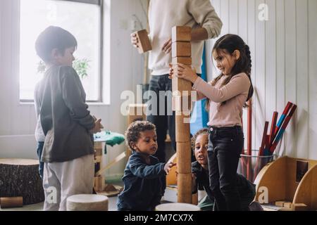 Weibliche und männliche Schüler stapeln hölzerne Spielzeugblöcke von männlichen Lehrern in Kindertagesstätten Stockfoto